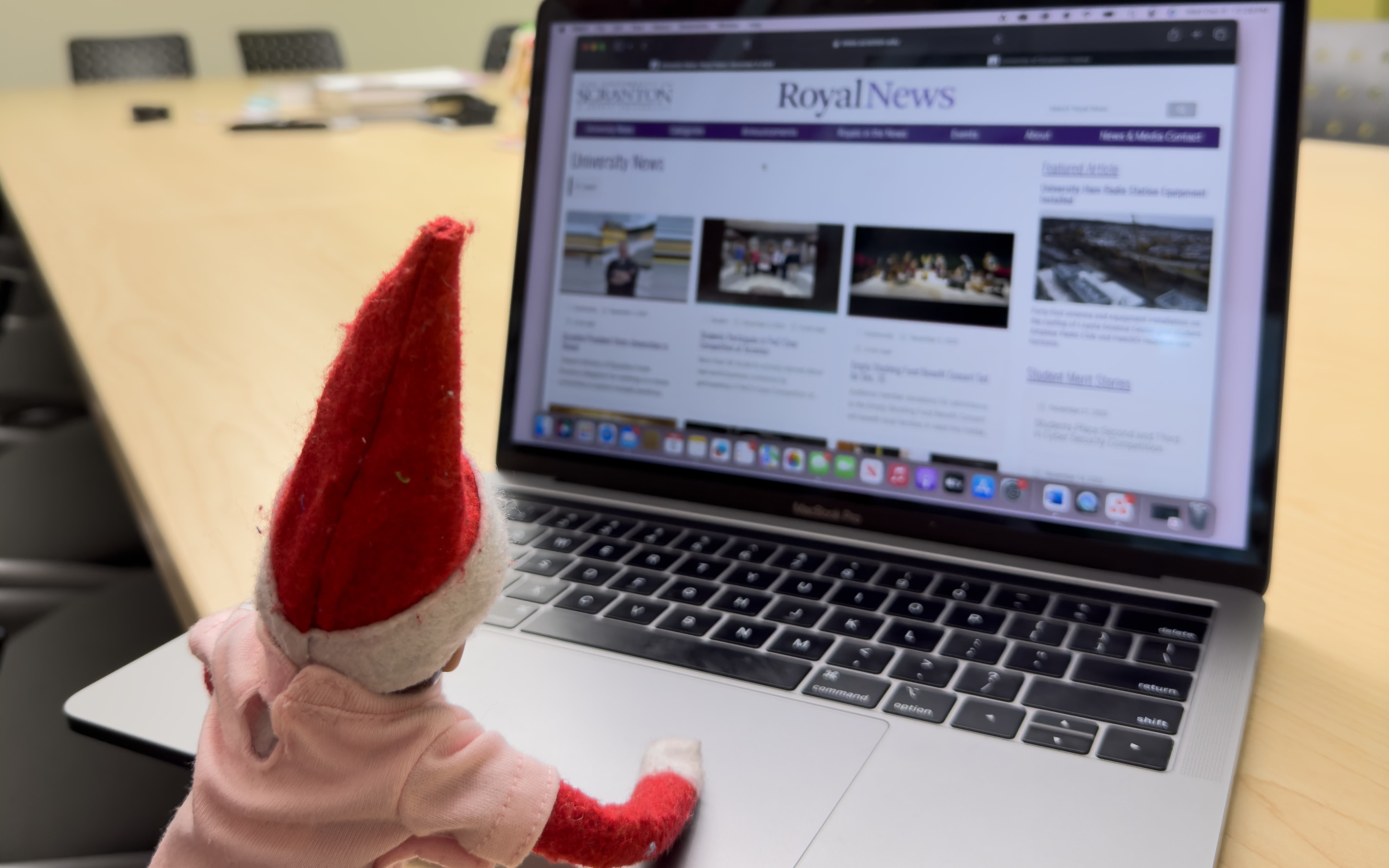 toy elf in holiday clothing sitting at a laptop with Royal News on its screen