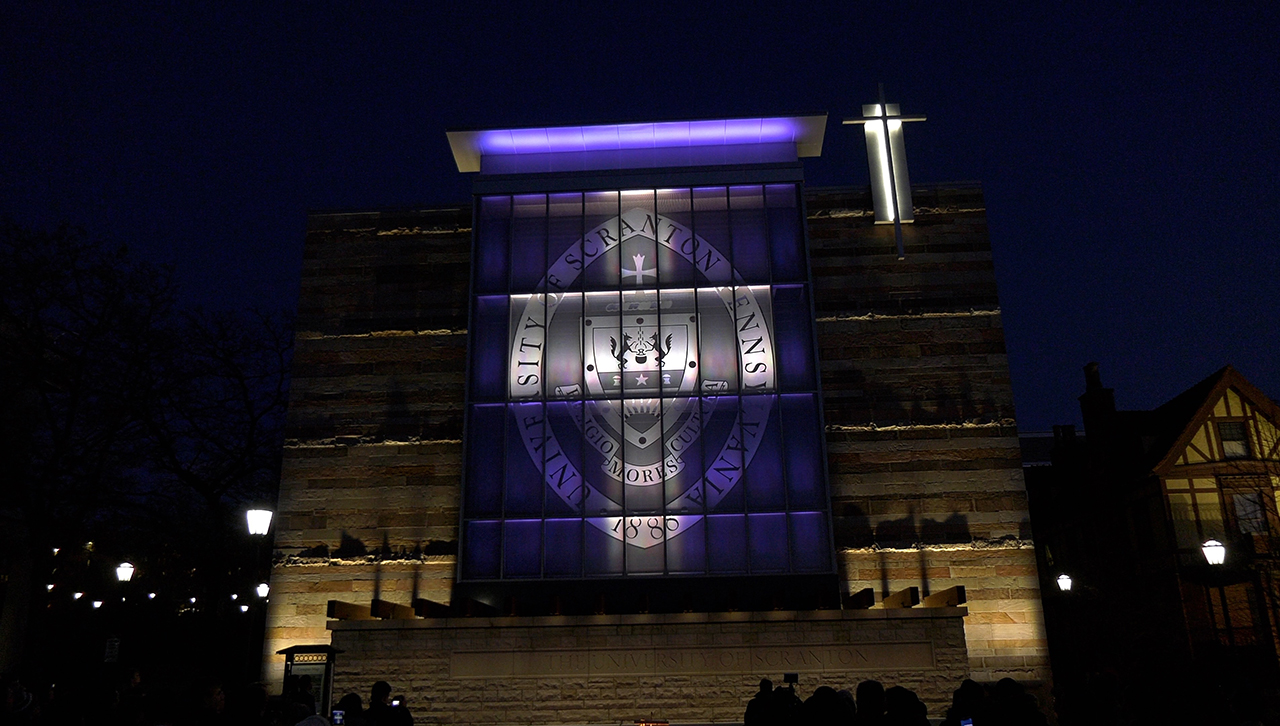 The Class of 2020 Gateway lit up at night to symbolize a large white cross against a purple background.