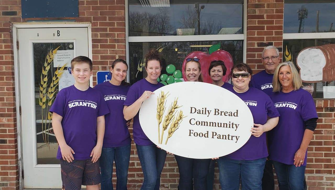 A group of 8 people wearing University of Scranton t-shirts gather around and/or hold a sign that reads "Daily Bread Community Food Pantry."
