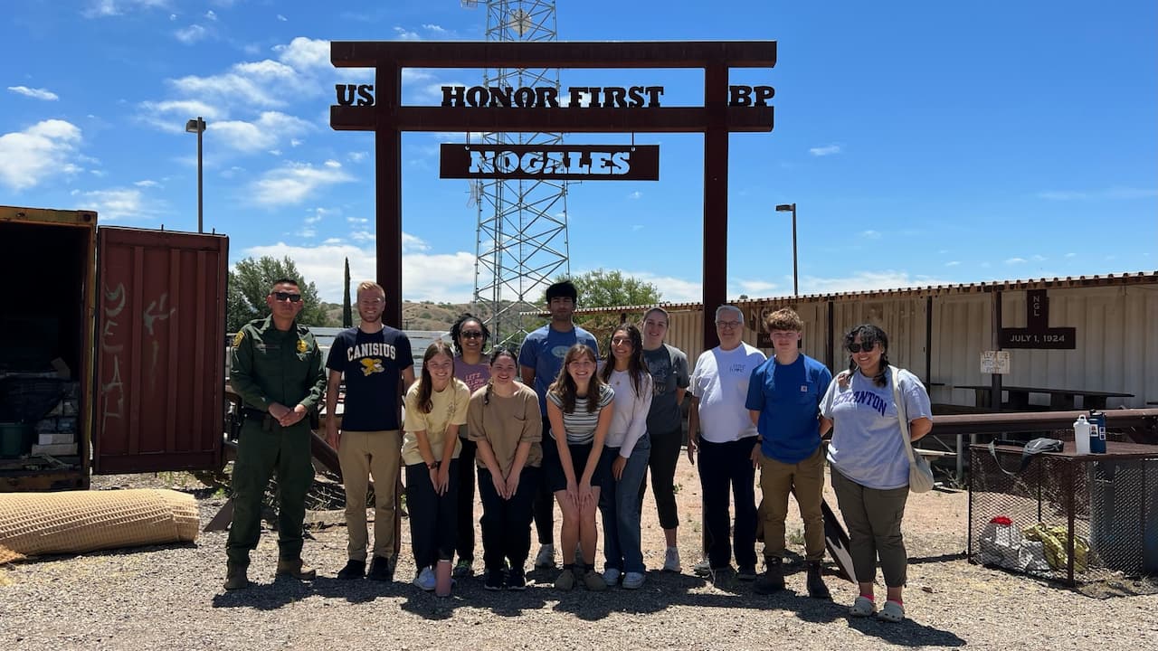 group of people standing at the Kino Border in Nogales, Mexico in front of an arch with a sign "US Honor First BP Nogales"