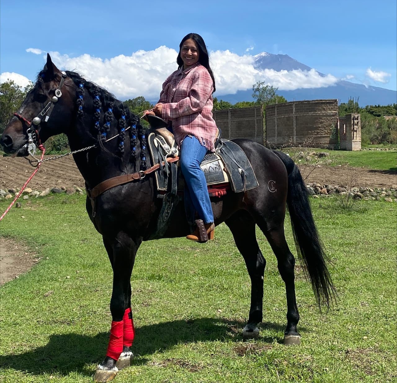 Yasmin Ramirez sitting atop a black horse, in front of clods adn mountainaous background in Mexico. 