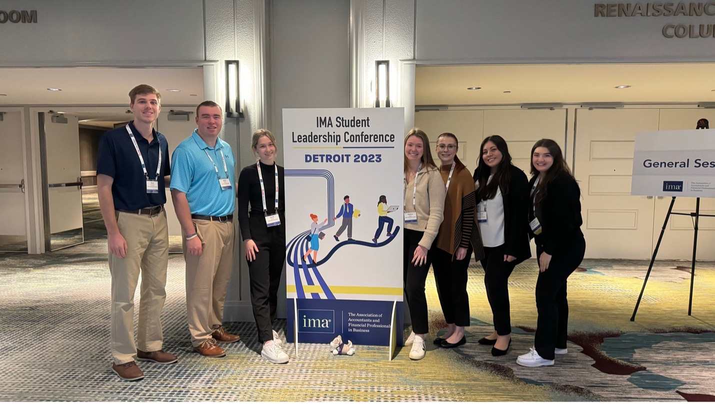 students and a professor standing in a convention center with a poster that includes the phrase Institute of Management Accountants Student Leadership Conference Detroit 2023