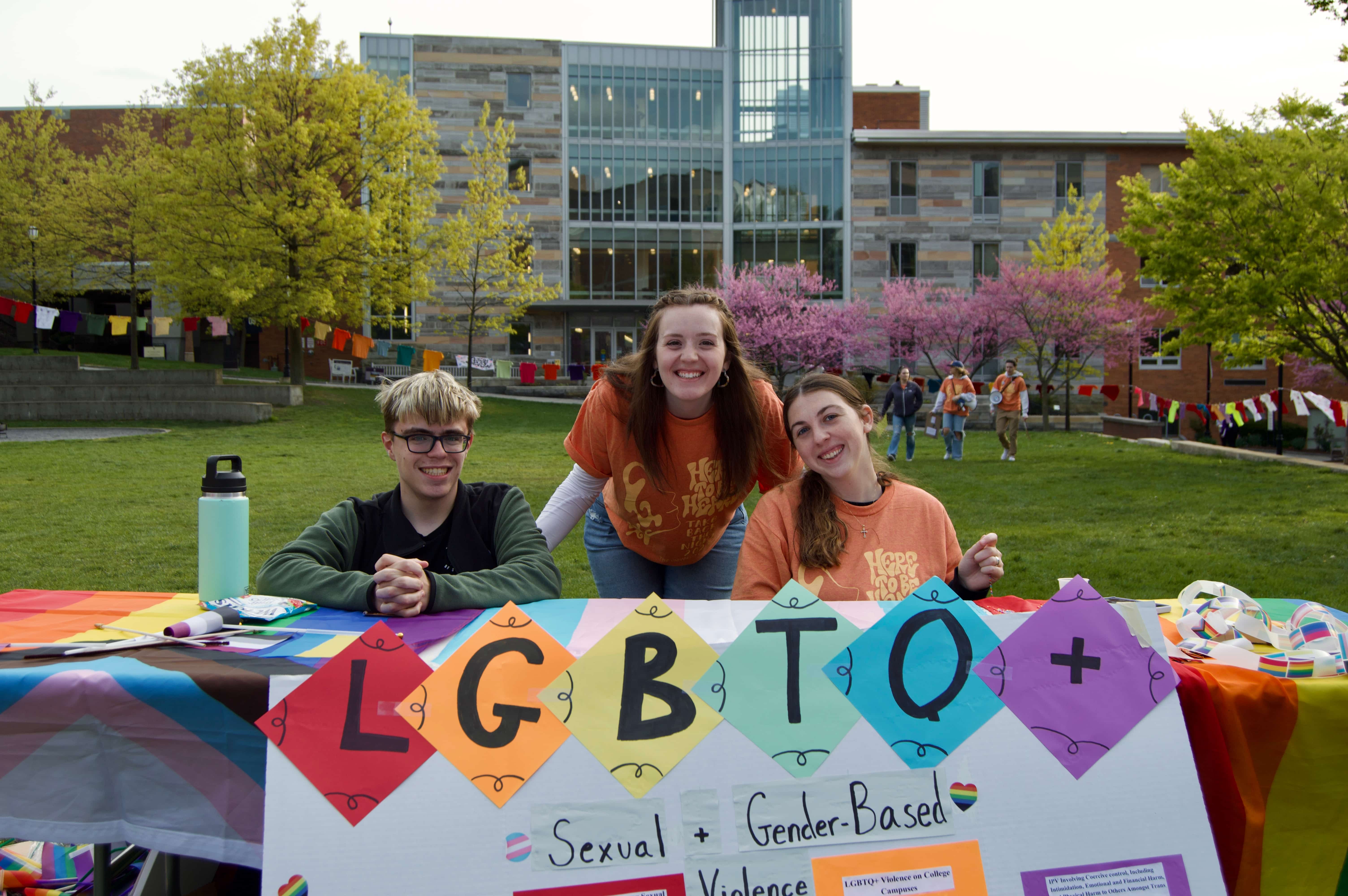 three people at a table on a lawn. In front of them a poster features the letter LGBTQ+