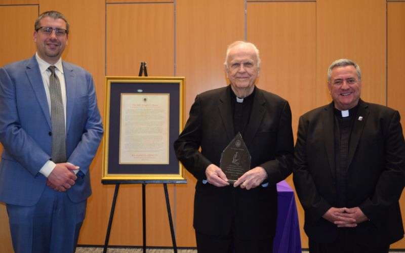 Three individuals pose for a photo during an award ceremony, with one recipient holding a plaque, while a framed document is displayed behind them.