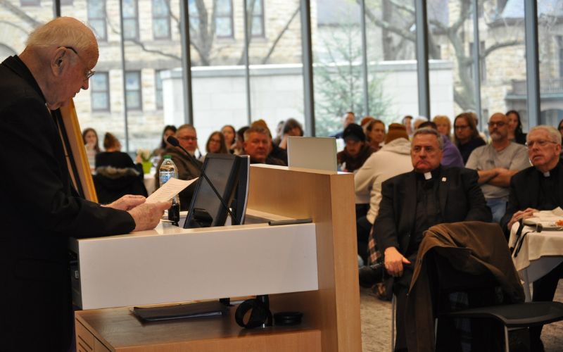 A speaker addresses an attentive audience in a modern conference setting. The room features large windows that allow natural light to illuminate the space.