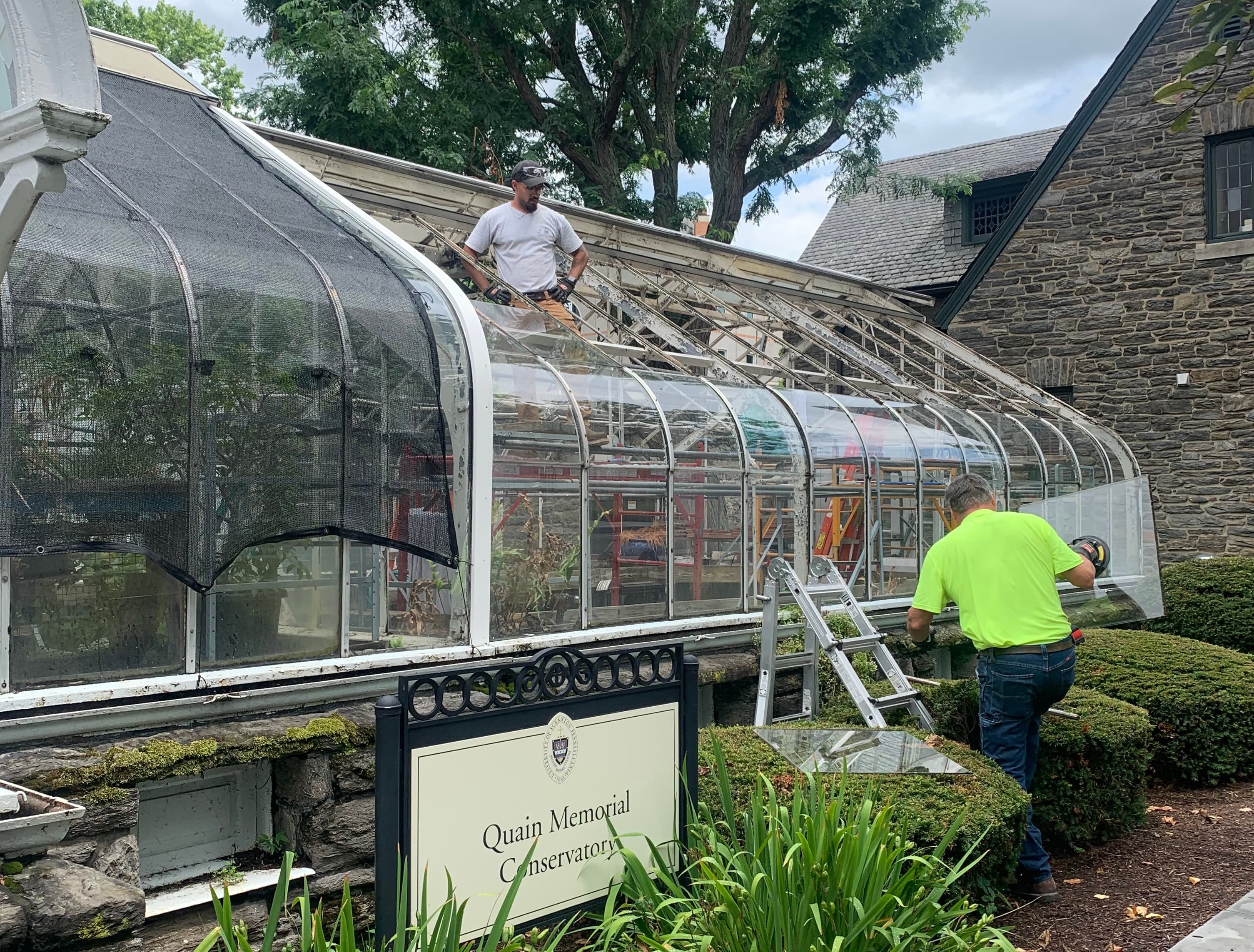 Two people working outside a greenhouse with a sign in front "Quain Memorial Conservatory"