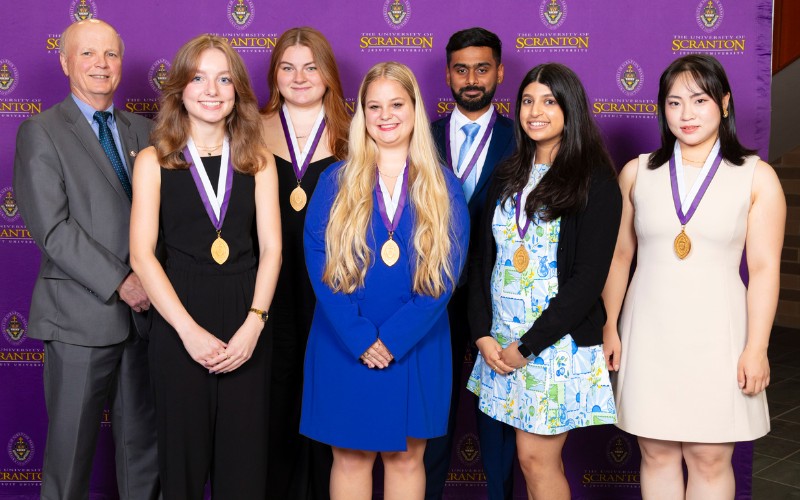 Group photo of students and faculty members at the University of Scranton award ceremony, featuring individuals wearing medals and standing against a purple backdrop with the university logo.
