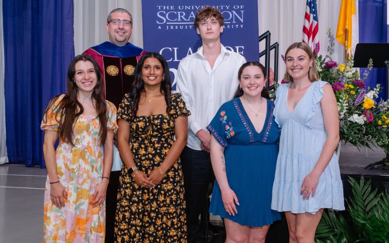 A group of graduates posing with a faculty member during a commencement ceremony at the University of Scranton.