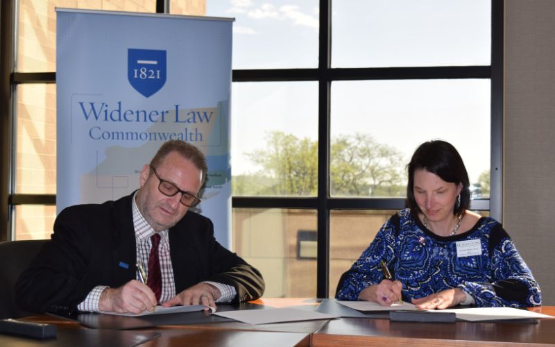 Two individuals signing documents at a table, with a banner for Widener Law Commonwealth in the background.