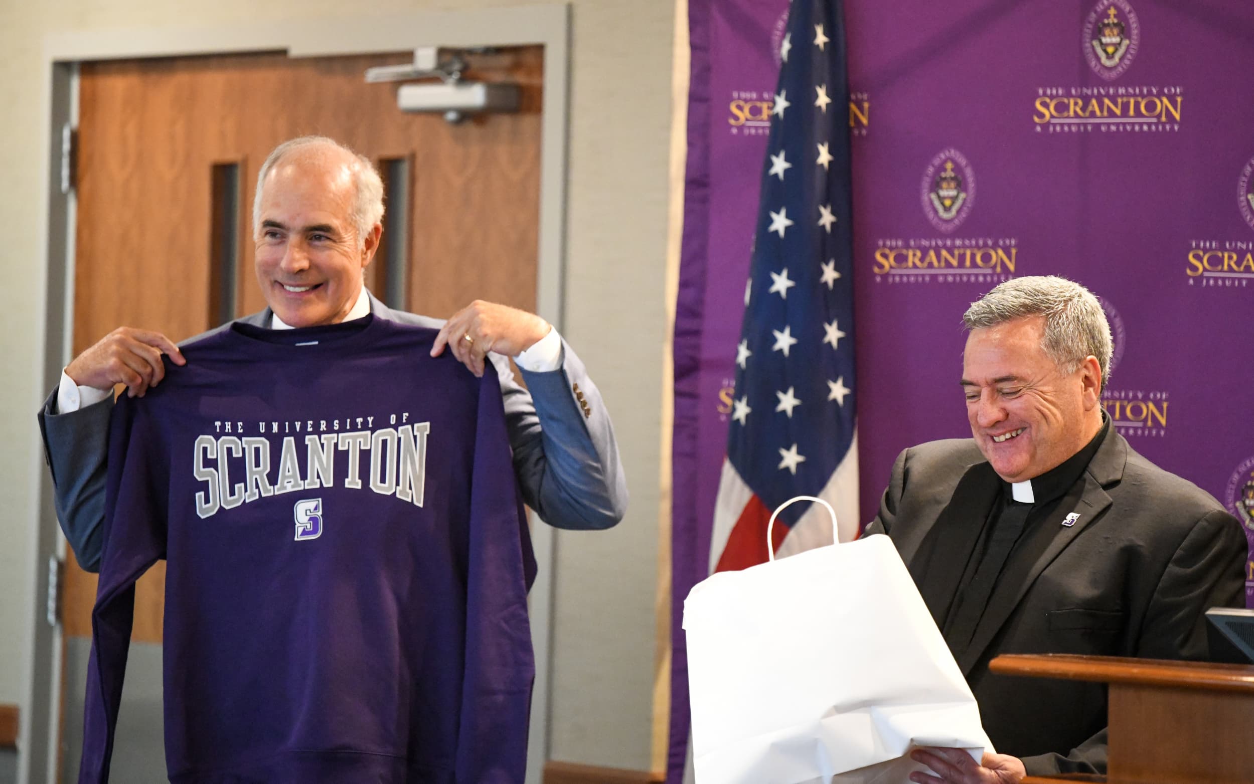 Two individuals stand at a news conference, one holding a University of Scranton T-shirt.