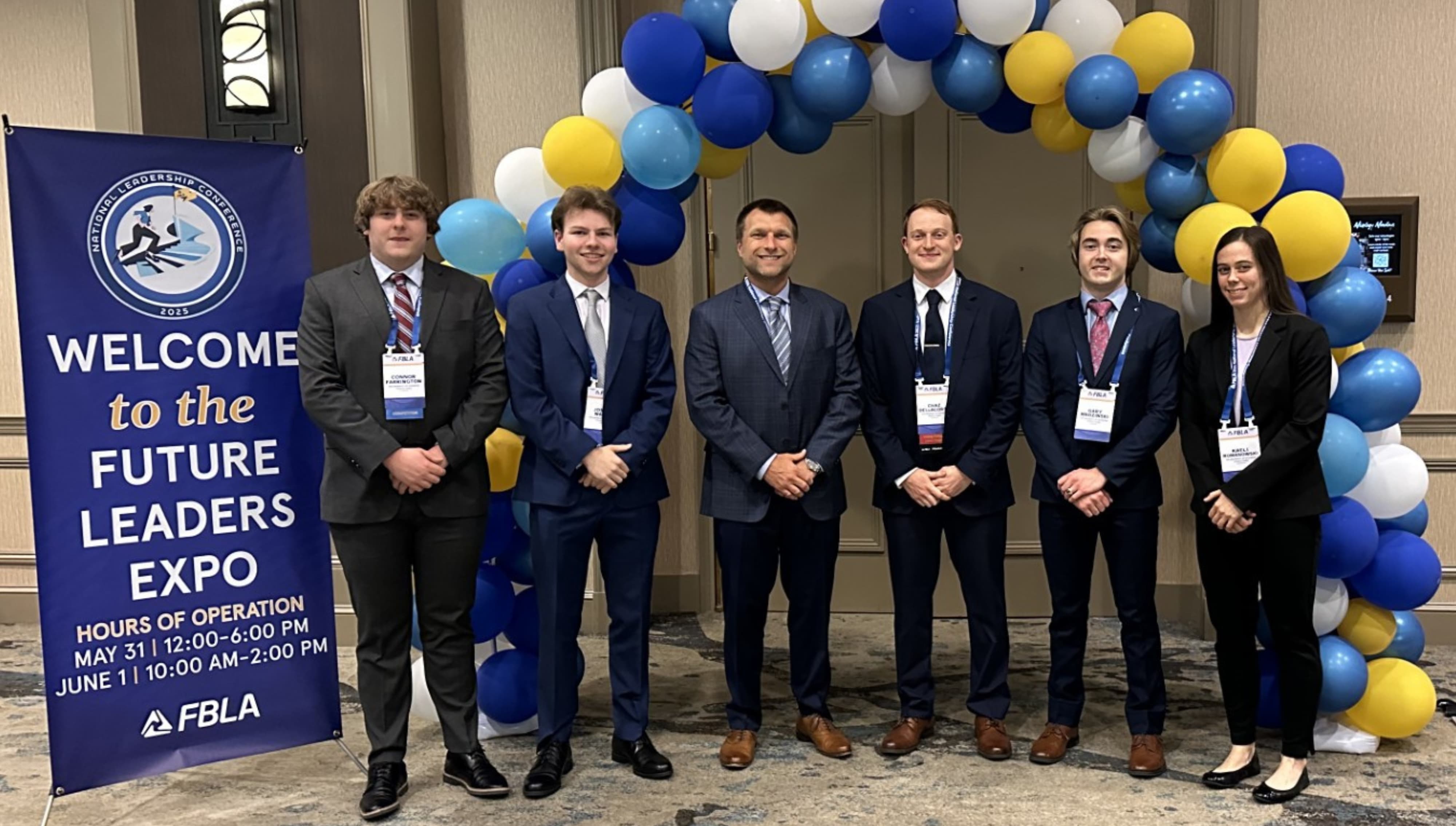 A group of six individuals posing in front of an arch made of blue and yellow balloons at an event.