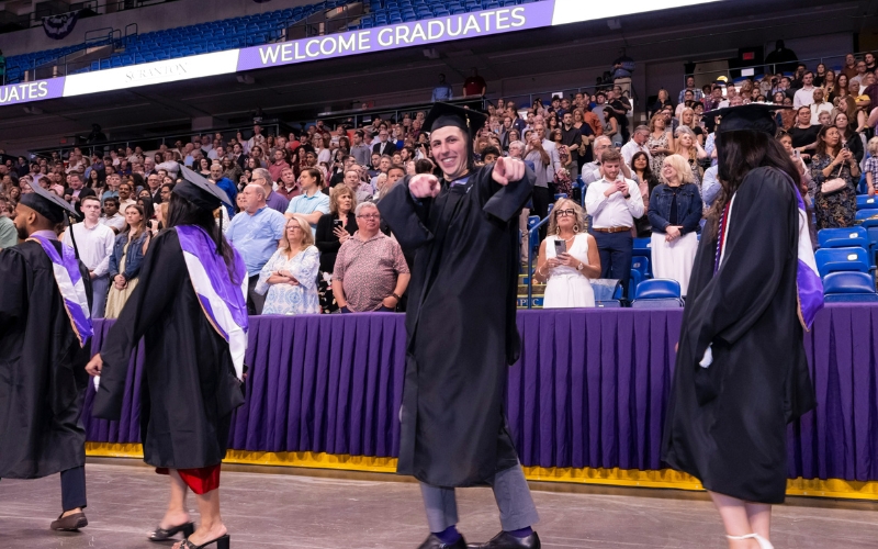 a smiling individual in a cap and gown points at the camera