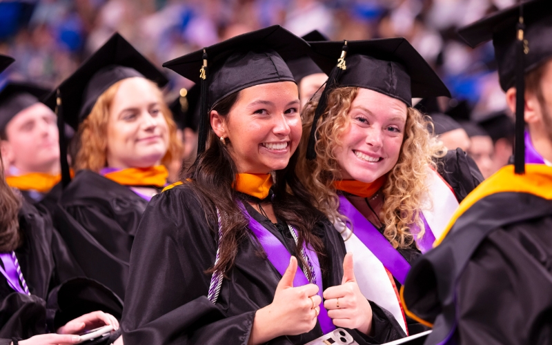 Two smiling graduates in caps and gowns giving thumbs up at a graduation ceremony.