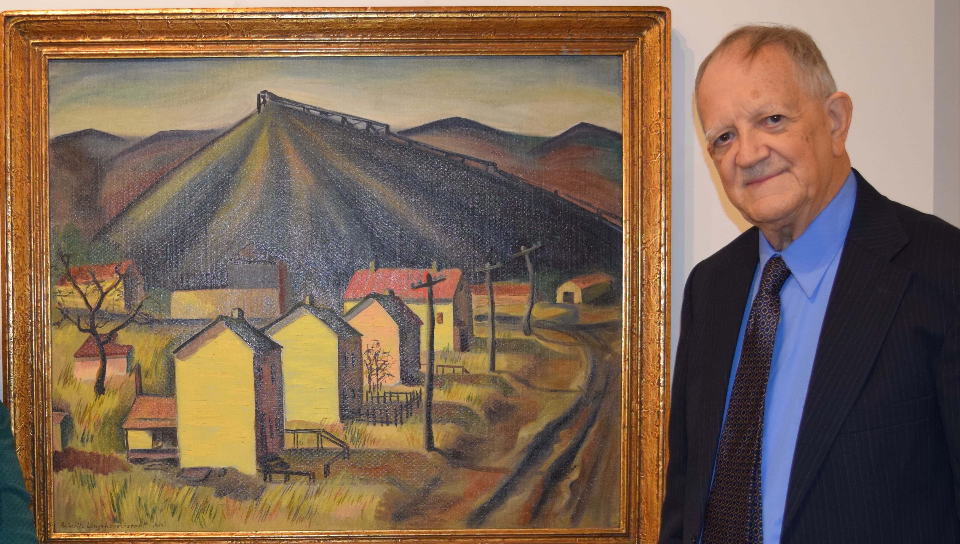 An individual stands beside a colorful painting depicting a rural landscape with yellow houses and rolling hills.