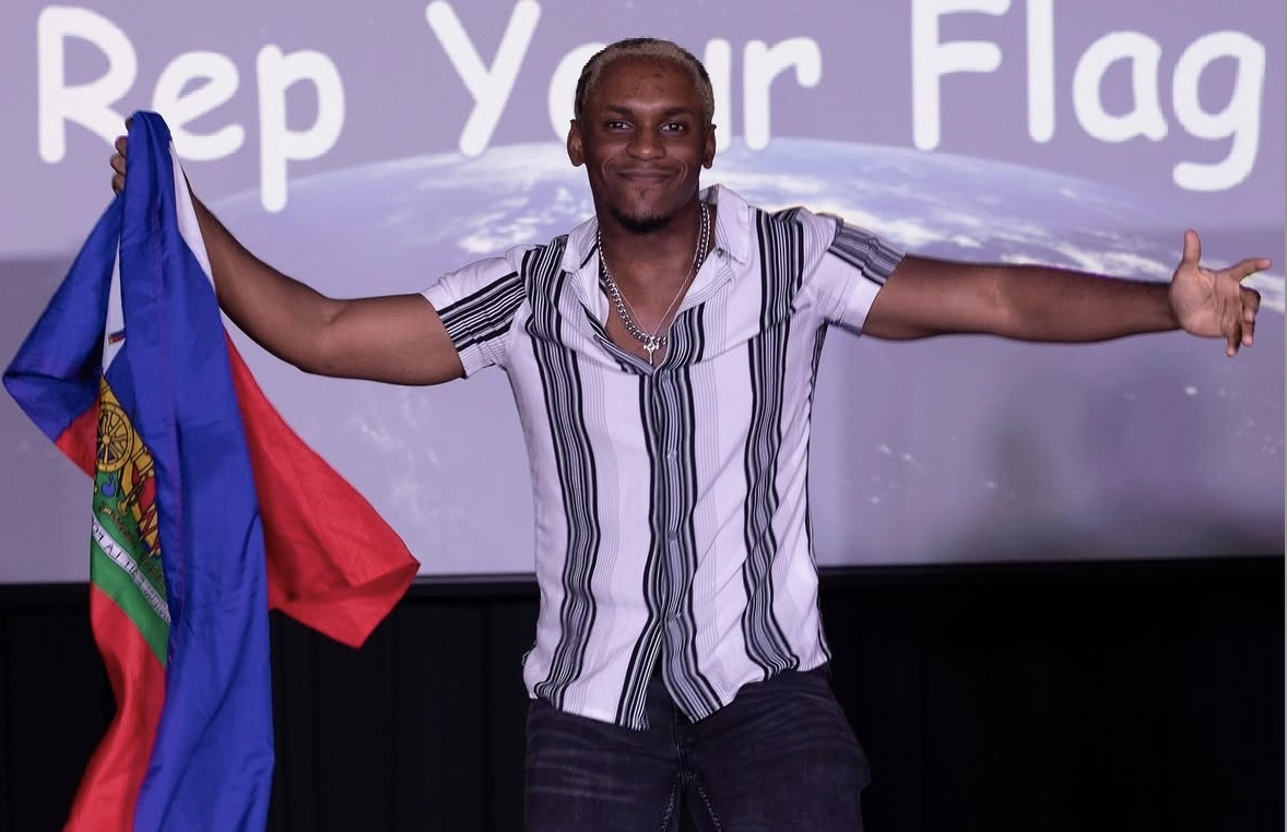 A young man walks on a runway carrying a flag. Behind him, the backdrop states "Rep Your Flag."