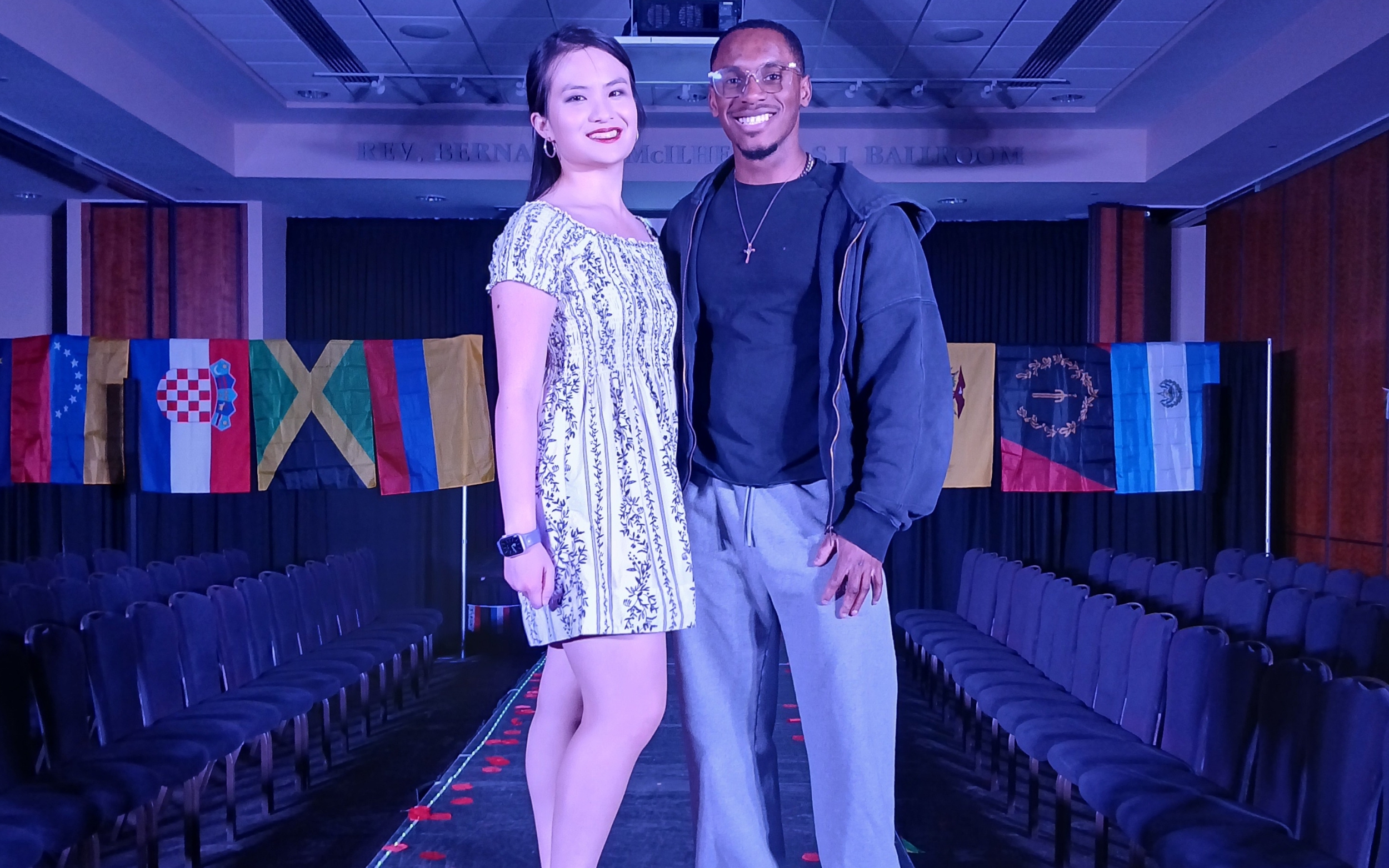 Two individuals pose on a fashion runway, with flags from various countries displayed in the background.