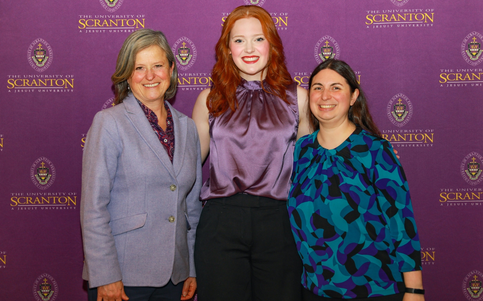 Three individuals pose together for a photo at The University of Scranton event, with a purple backdrop featuring the university's logo.