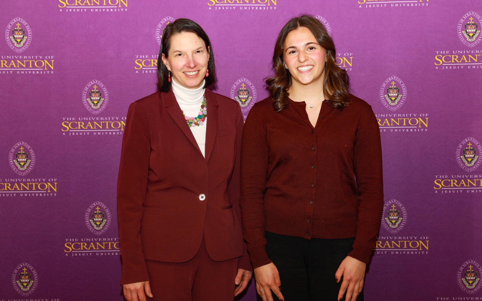 Two individuals pose for a photo together against a purple backdrop featuring the University of Scranton logo.