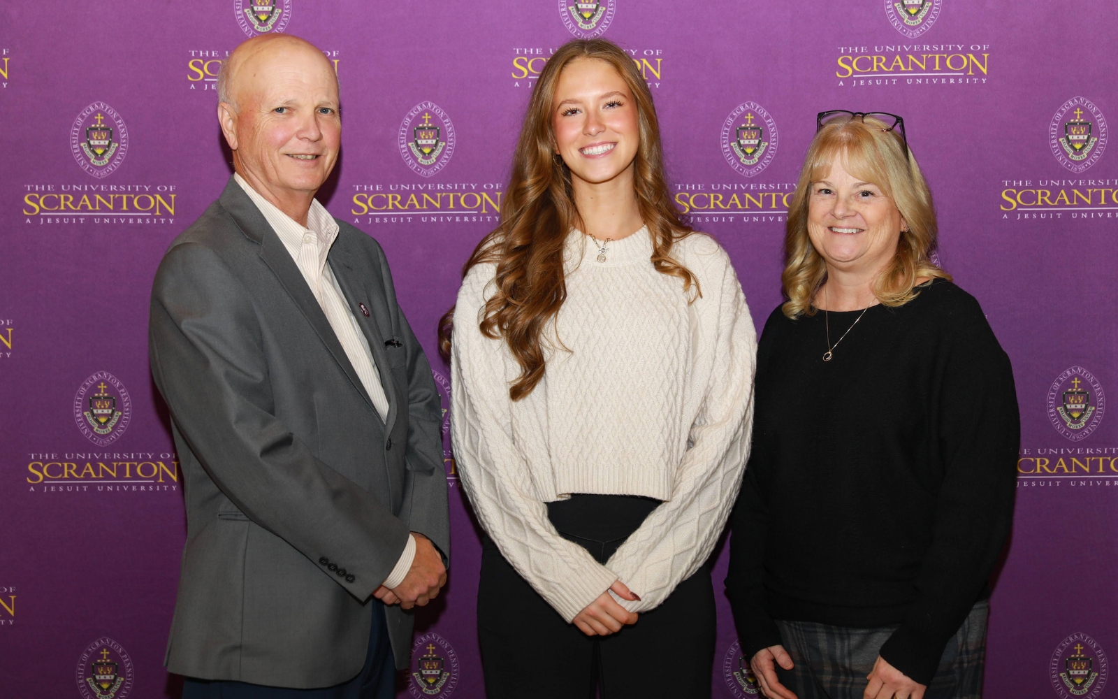 Three individuals pose together in front of a University of Scranton backdrop.