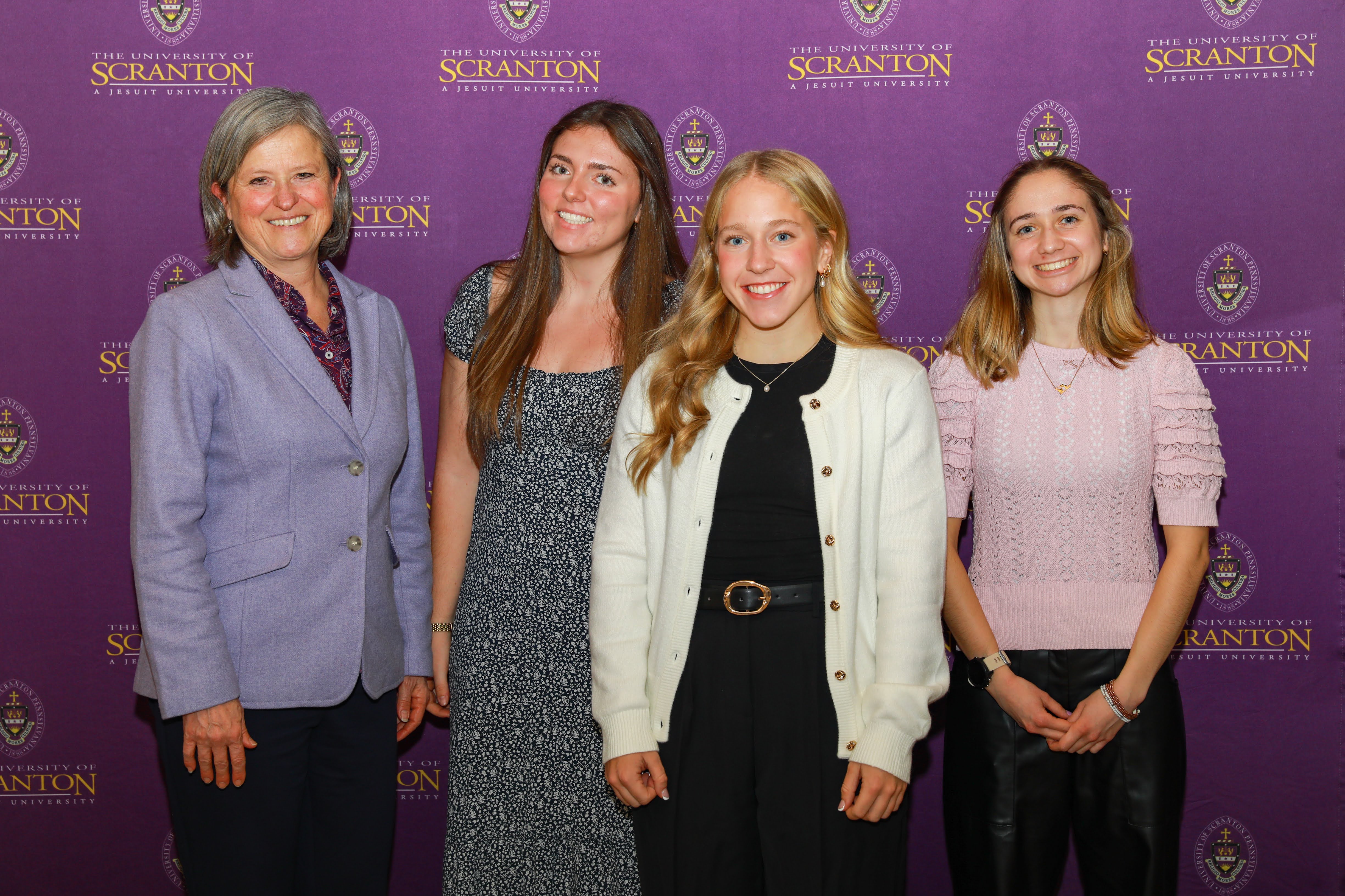 Four individuals pose for a photo at an event hosted by the University of Scranton, smiling in front of a purple backdrop featuring the university logo.