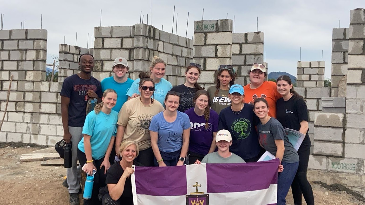 Fourteen University of Scranton occupational therapy students and their faculty leader stand in front of the progressing house build in San Juan de la Maguana, Dominican Republic.