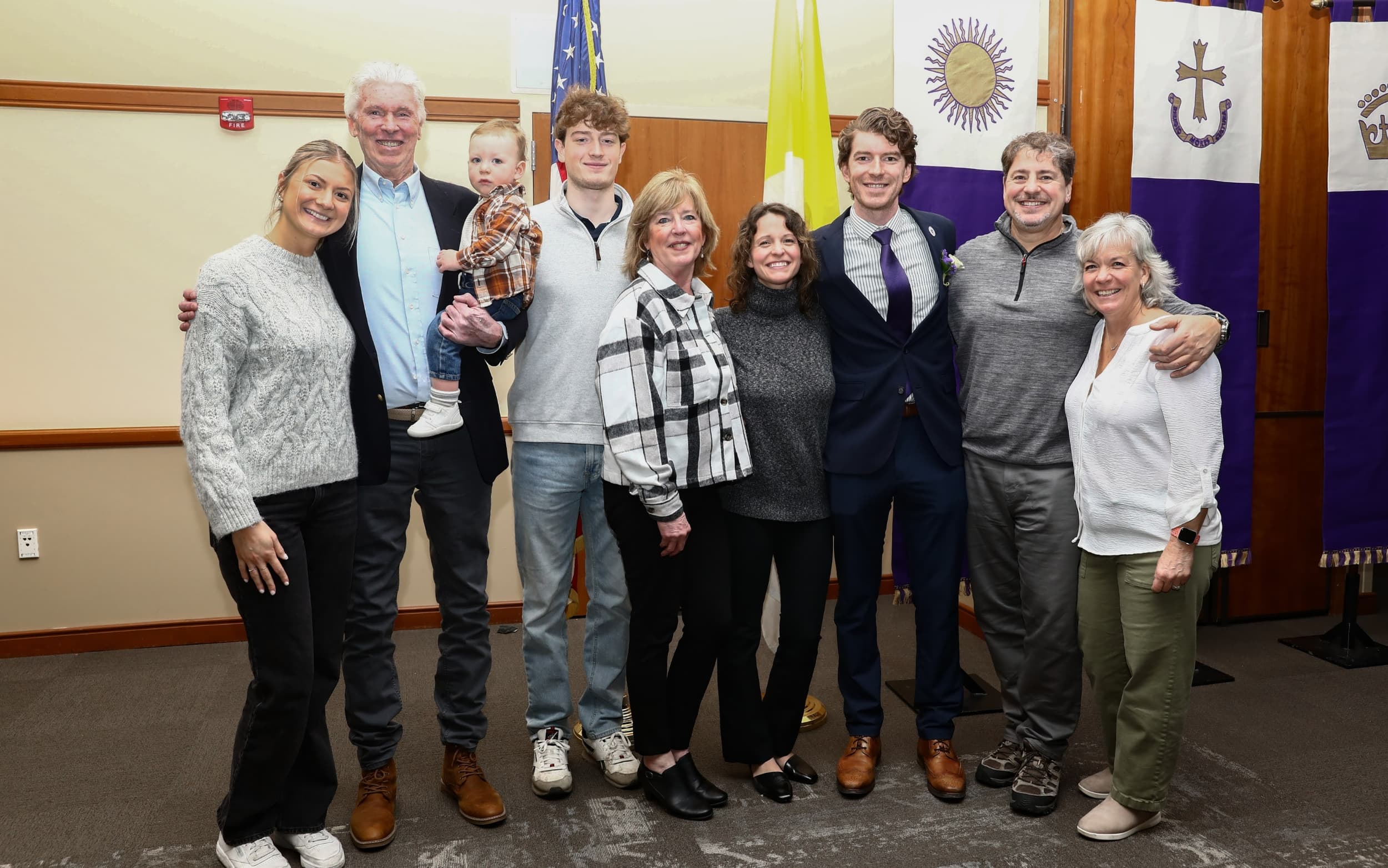 Group of people posing indoors for a family photo, with U.S. and purple ceremonial flags in the background.