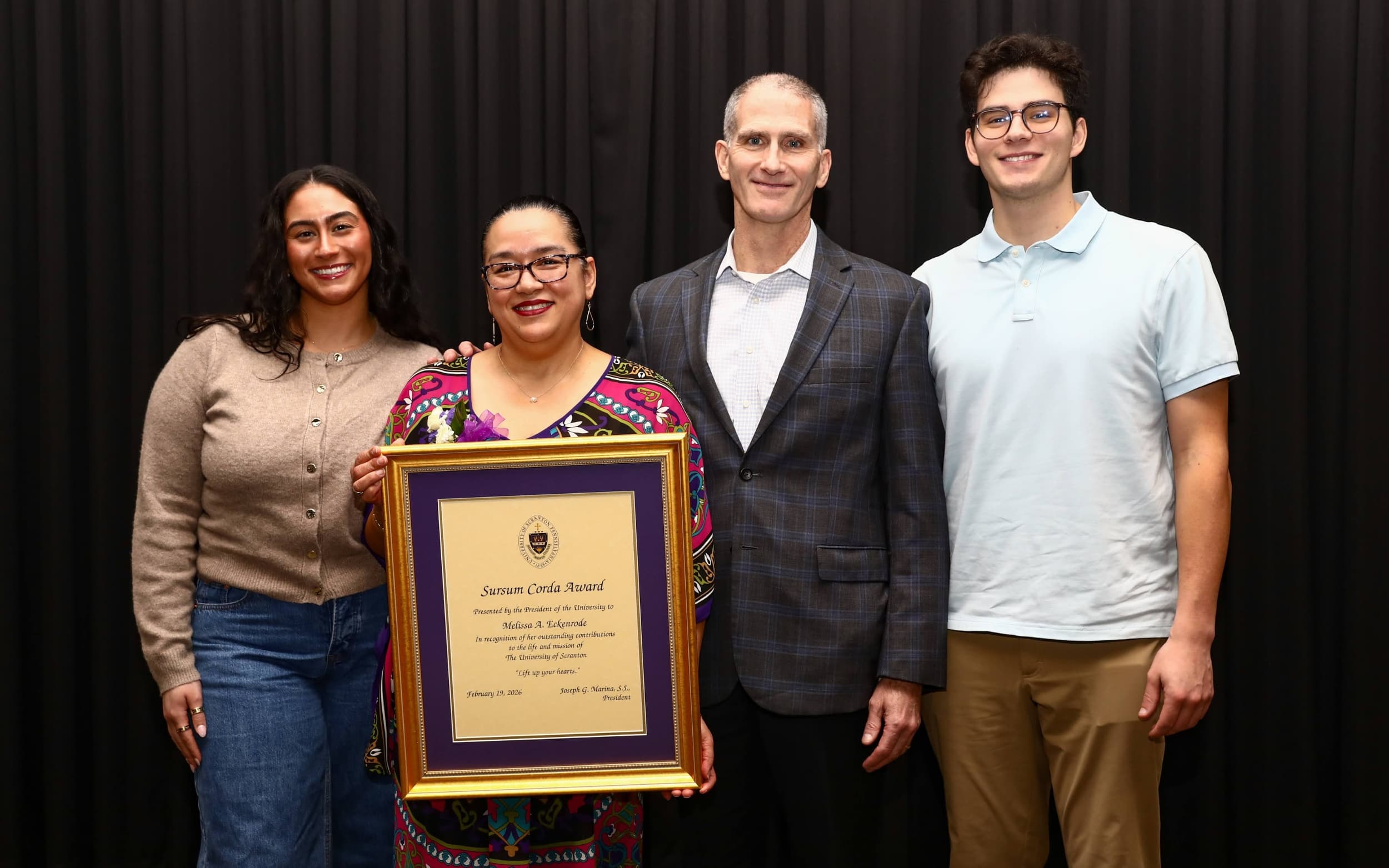 Four people posing at an awards event, with one holding a framed certificate in front of a black curtain backdrop.