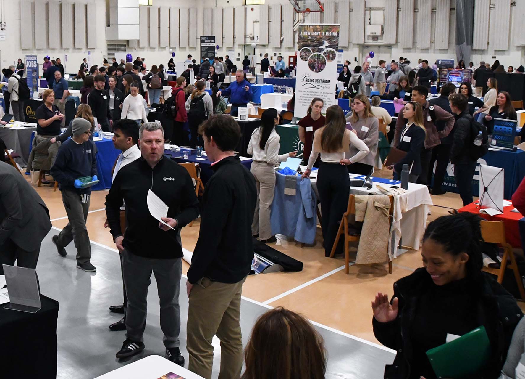 overview photo of auditorium space featuring a crowd of adults in conversation at vendor tables decorated with purple and white balloons