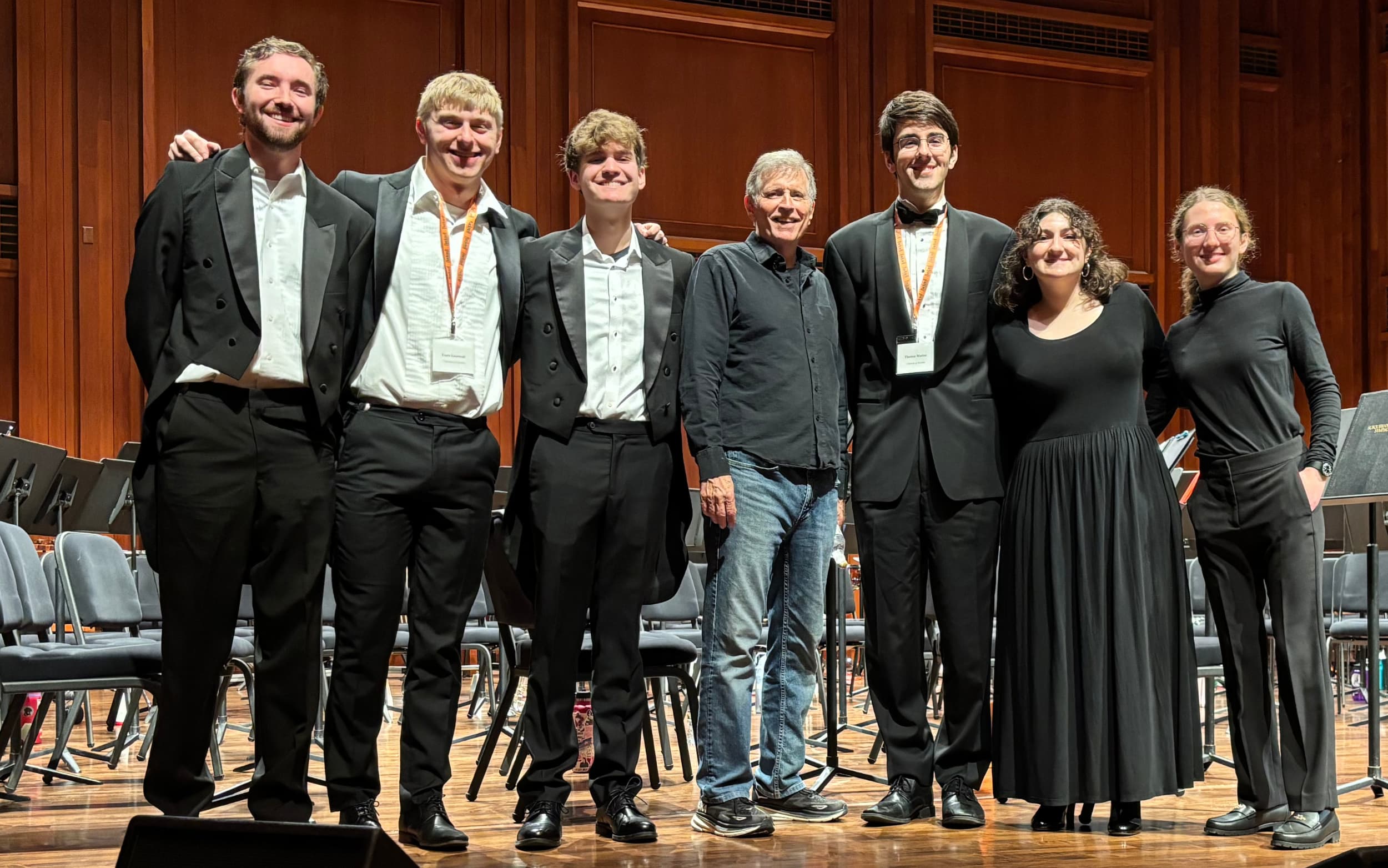 Group of musicians and a conductor pose together on a concert hall stage with chairs and music stands behind them.