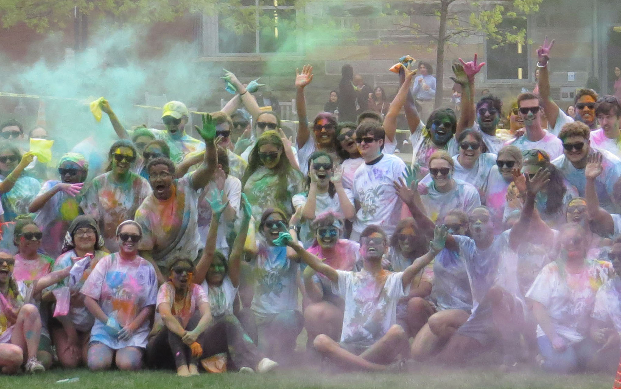 crowd of people in a green space, wearing white T-shirts covered in colored powder