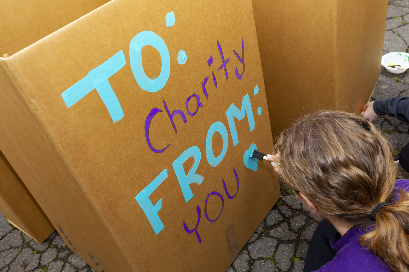 Shown is a person painting a sign on a large cardboard box used to collect donations: "To: Charity From: You