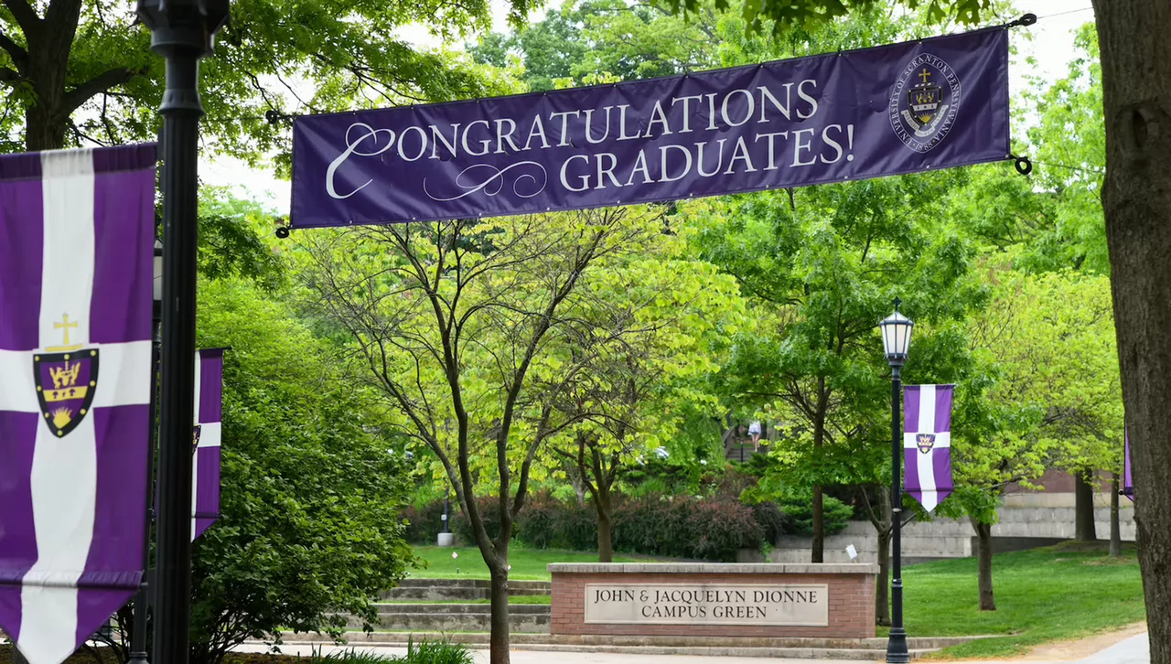 Purple campus banner reading “Congratulations Graduates!” hanging between lampposts amid leafy green trees.