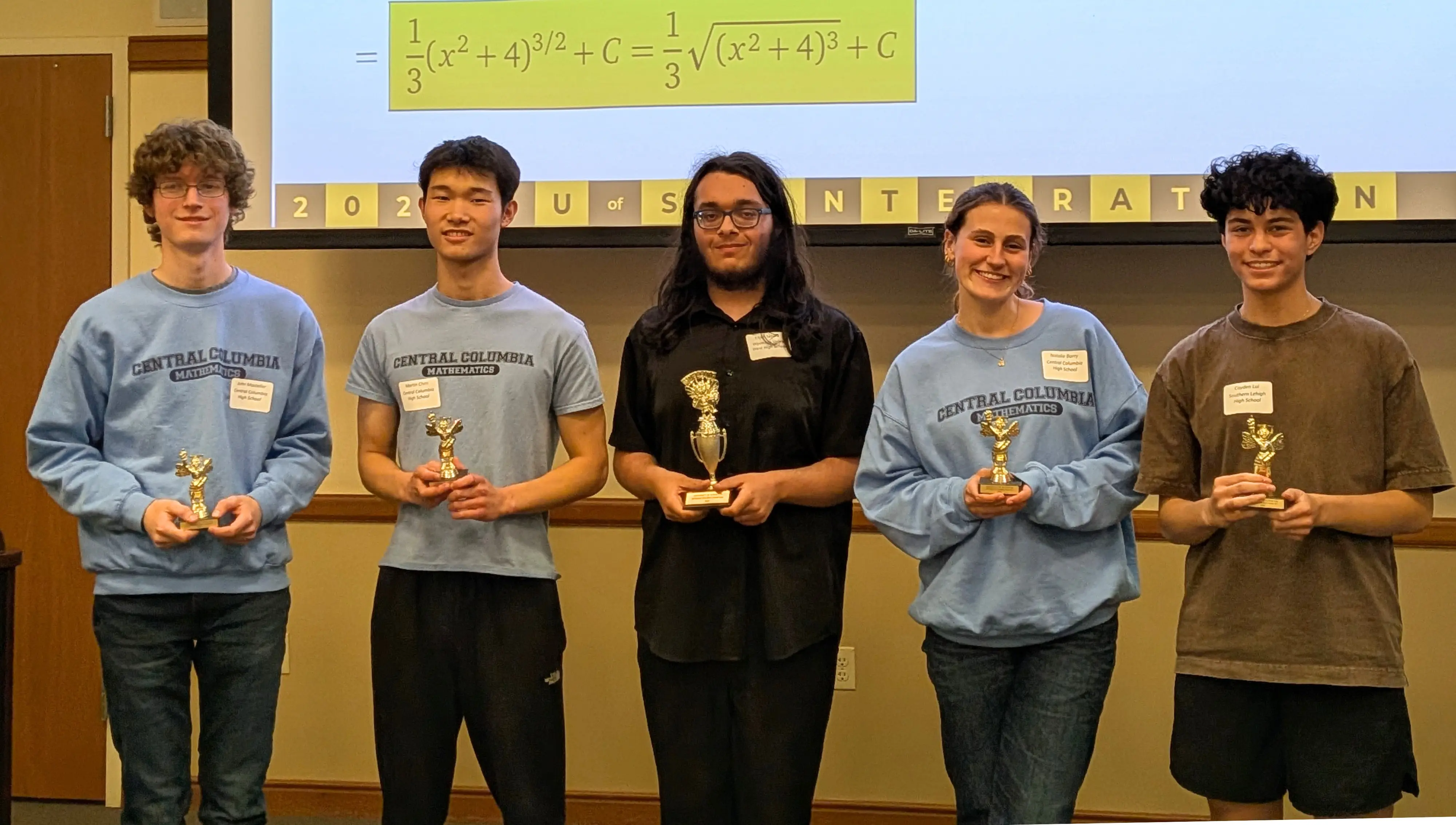 Five students pose indoors holding trophies, with a projected math equation displayed above them.