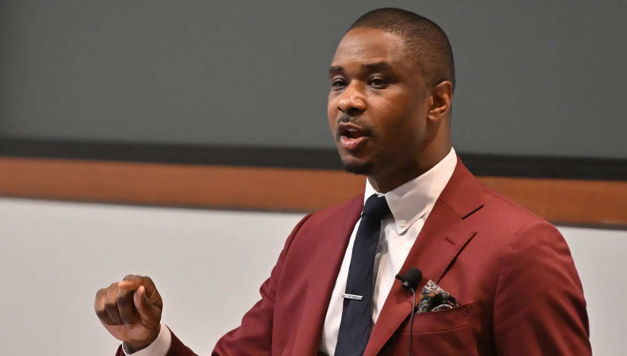 Individual in a suit speaking during a presentation, gesturing with one hand.