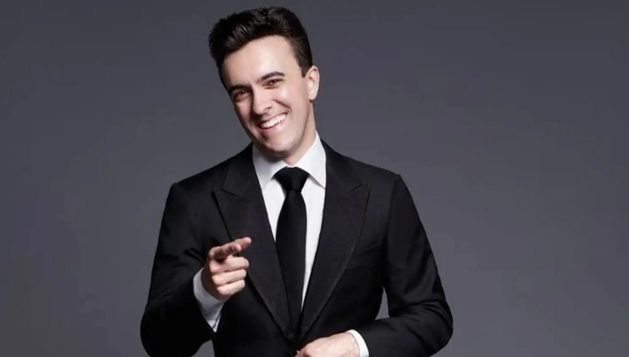Smiling individual in a black suit and tie pointing toward the camera against a gray background.