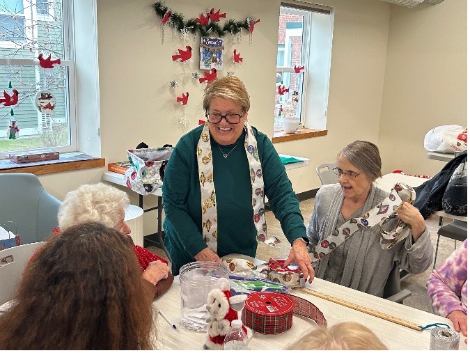 in a room adorned with winter holiday decorations, a smiling woman helps wrap packages with older adults seated around a table