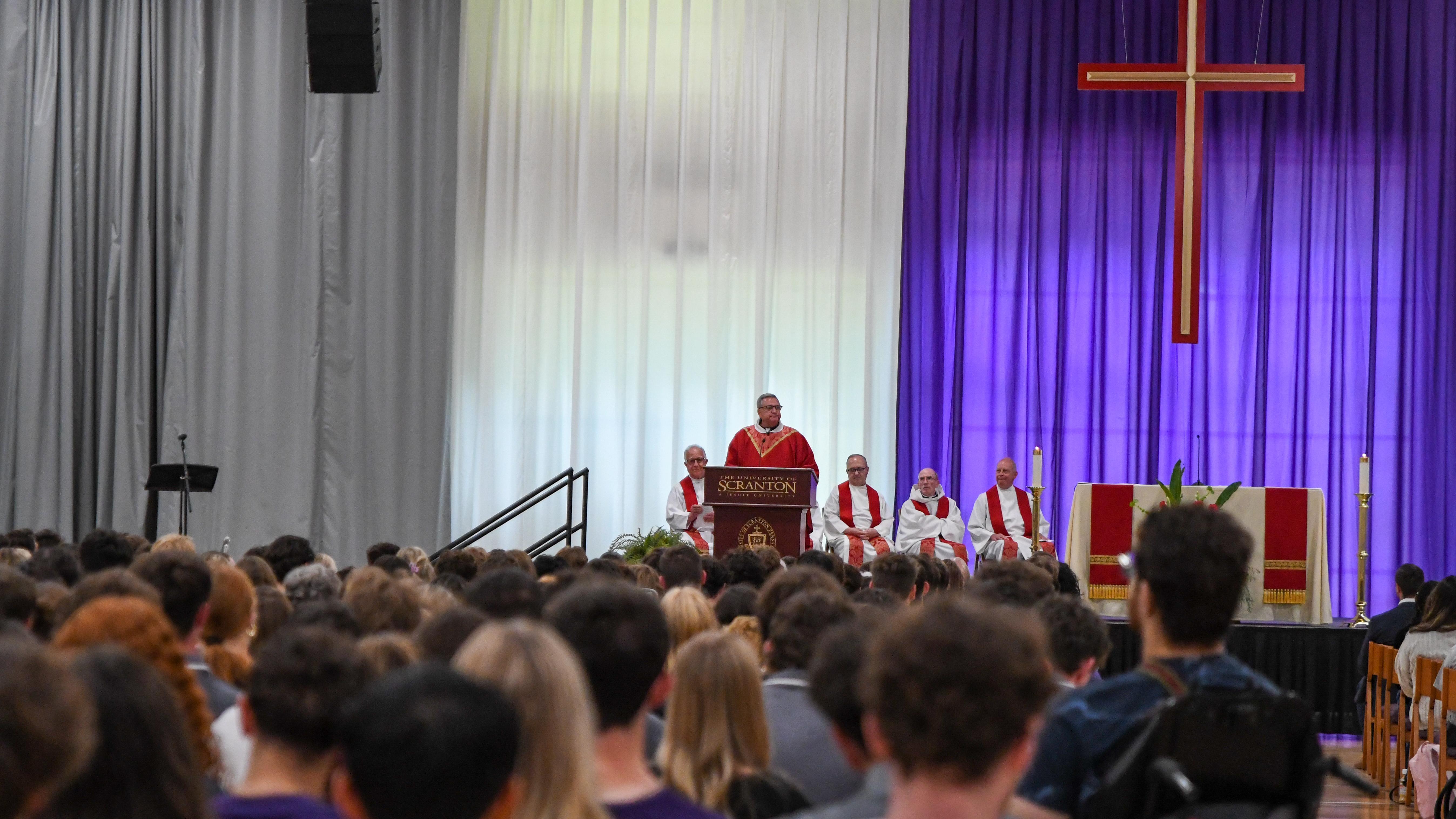 crowd seated at a liturgy with celebrant of the Mass wearing red vestments is speaking at a lectern