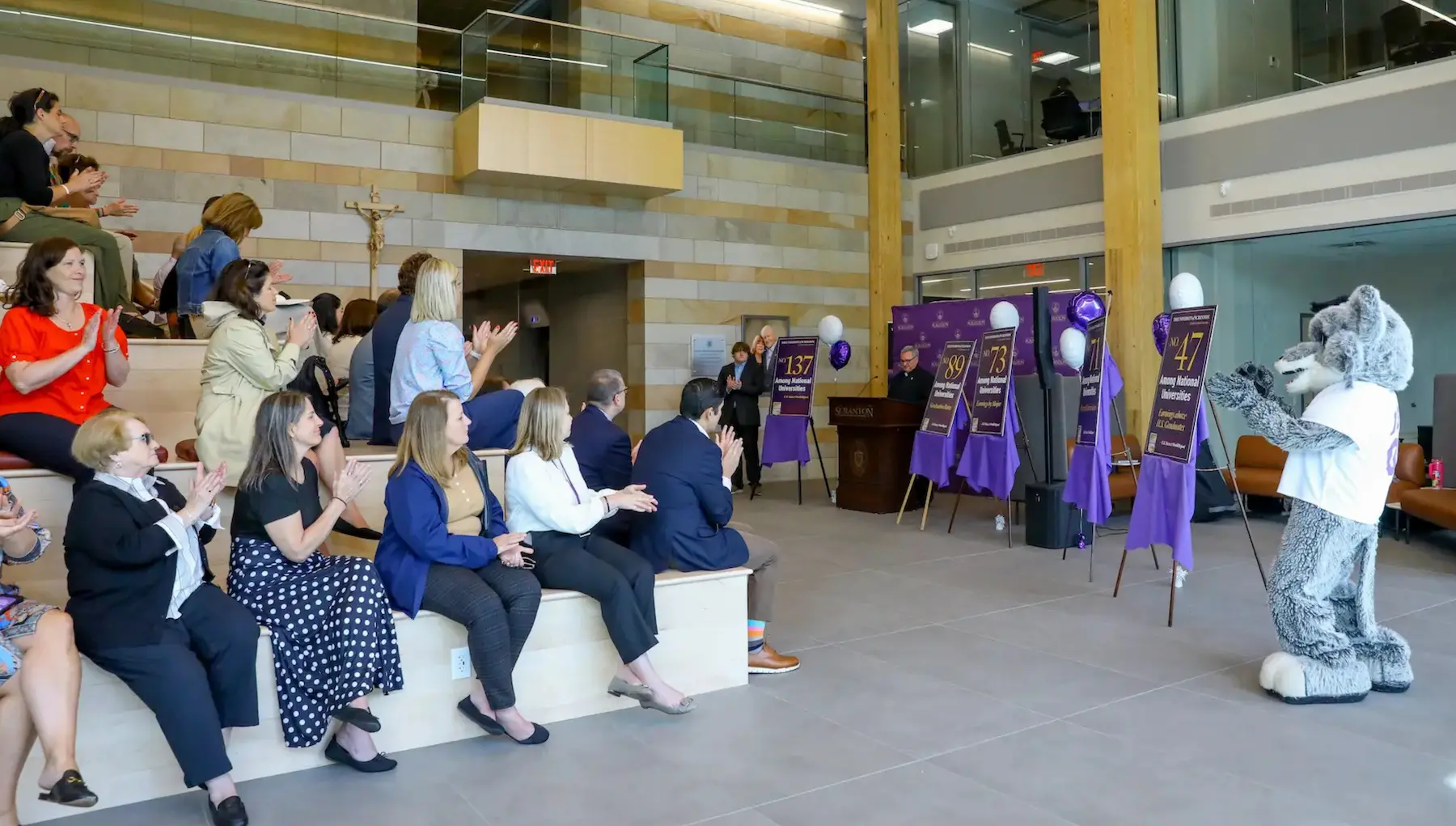 Audience applauds at an indoor university event as a mascot claps near purple balloons and ranking display posters.