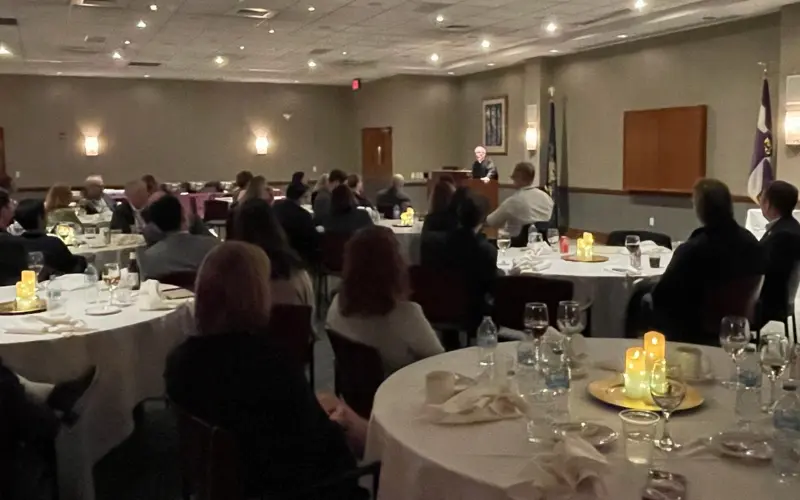 Audience seated at round, candlelit banquet tables listens to a speaker at a podium in a hotel conference room.