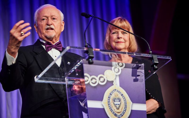 Two speakers stand behind a clear podium on a stage, with the man gesturing as he addresses an audience and a large medallion graphic displayed on the front.